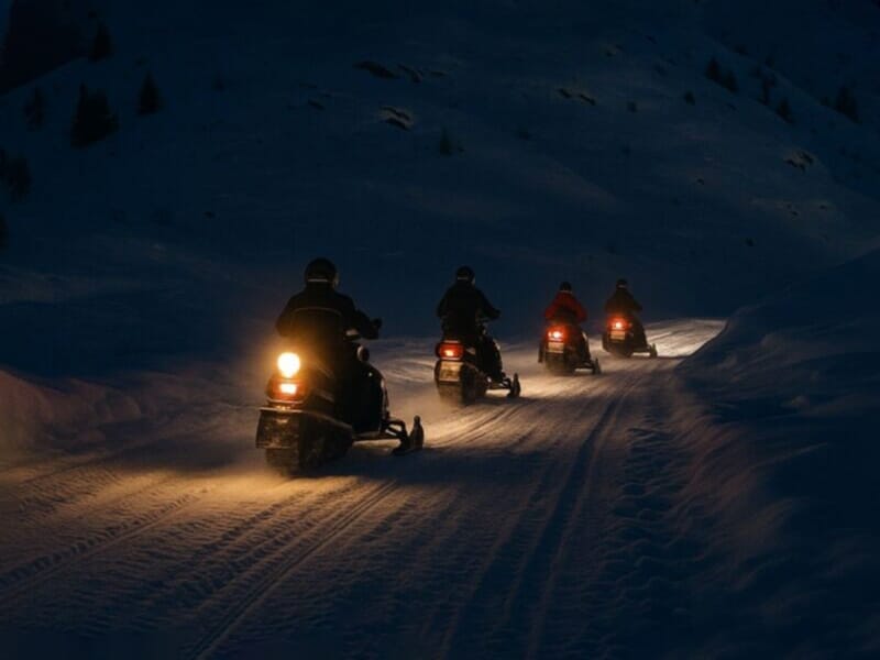 Groupe de motoneiges avec phares allumés roulant de nuit sur un sentier enneigé en montagne.