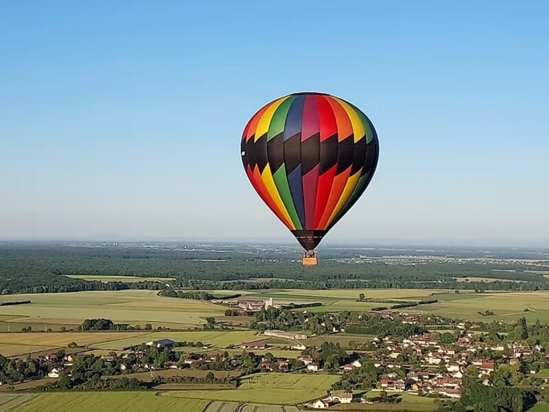 Montgolfière multicolore survolant un village et des champs verts sous un ciel bleu clair