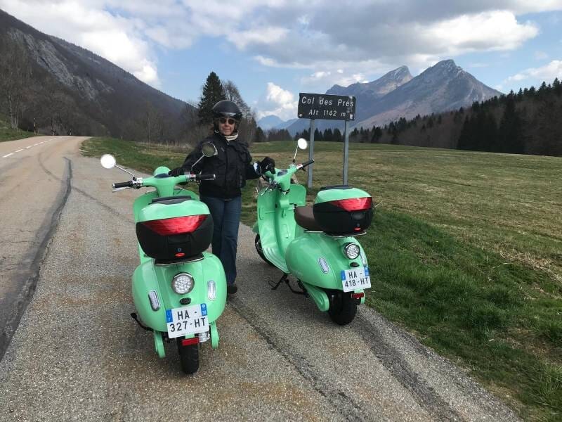 Personne en tenue de moto debout entre deux scooters verts sur une route de montagne au Col des Prés.