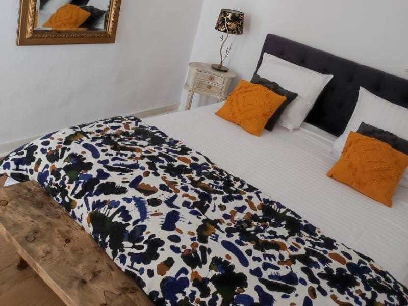 Chambre avec lit double, coussins orange, couverture à motifs et table de chevet avec lampe.