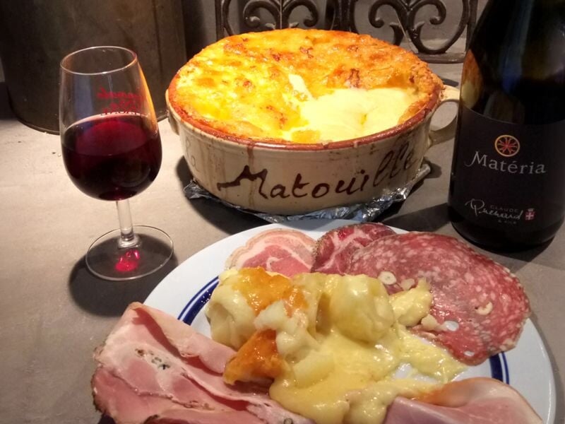 Assiette de charcuterie variée avec raclette fondue, verre de vin rouge et bouteille de vin Matéria sur table
