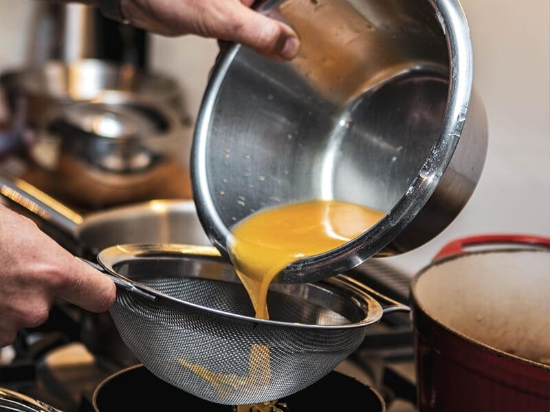 Main versant un liquide jaune à travers un tamis au-dessus d'une casserole lors d'un cours de pâtisserie à Lille.