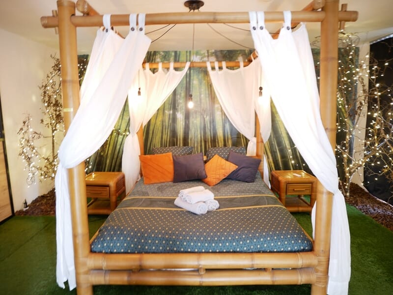 Lit à baldaquin en bambou avec rideaux blancs, coussins colorés et serviettes pliées lors d'une nuit en Love Room près de Châteauroux.