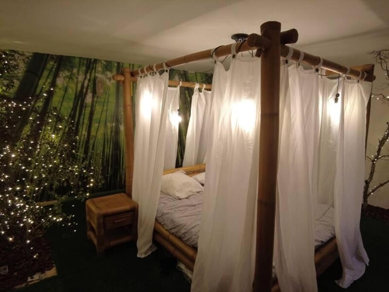 Lit à baldaquin en bambou avec rideaux blancs tirés, table de chevet en bois lors d'une nuit en Love Room près de Châteauroux.
