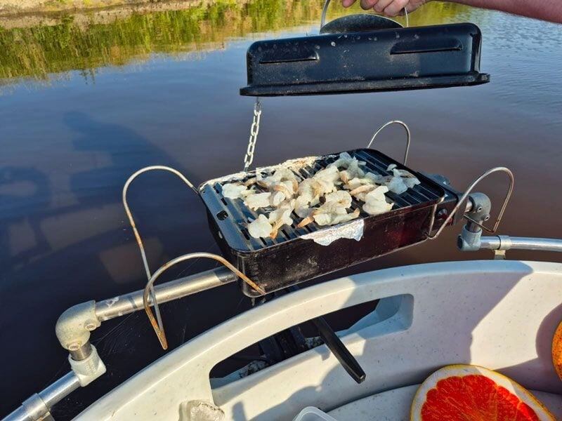 Barbecue portable sur un bateau avec des morceaux de viande en train de cuire au-dessus de l'eau calme.