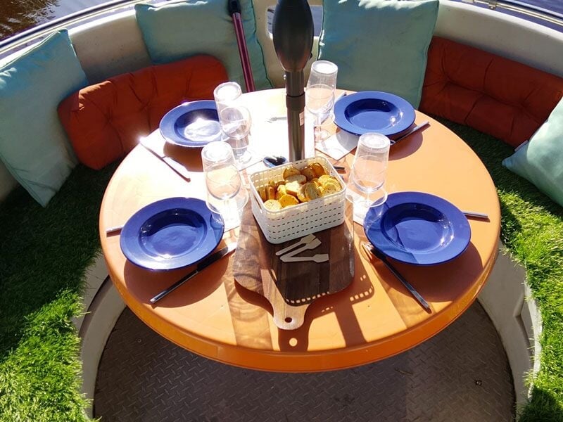 Table ronde sur un bateau avec assiettes bleues, verres et panier de pain pour un repas en plein air.