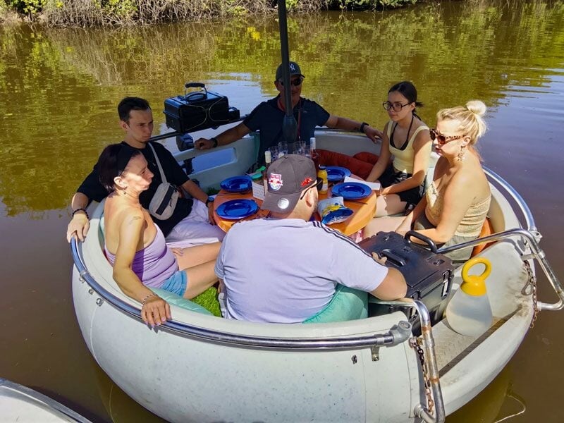 Six personnes assises dans un bateau rond sur l'eau, discutant et partageant un repas ensemble.