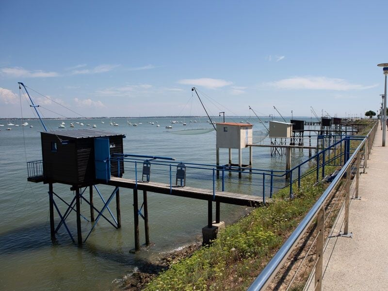 Cabane de pêche sur pilotis avec ponton en bois, vue sur la mer et plusieurs bateaux à l'horizon sous un ciel clair.