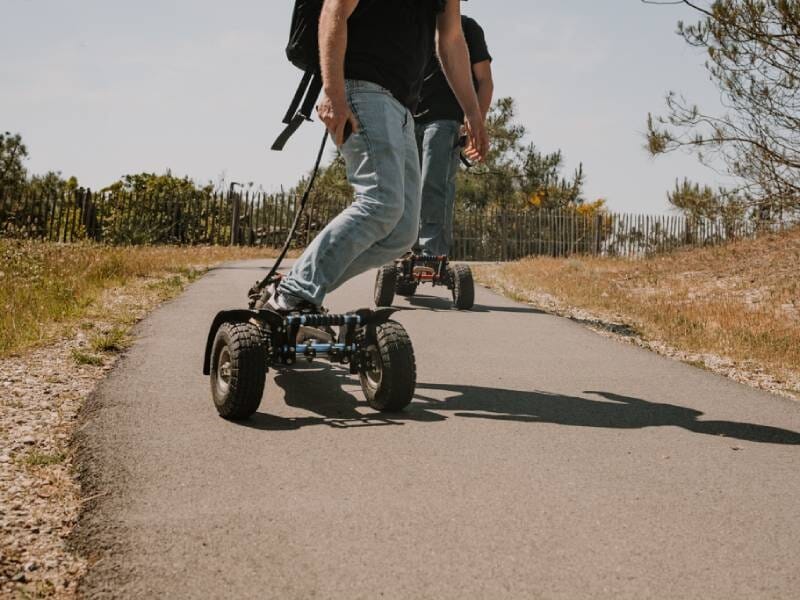 Personne en jean et t-shirt noir roulant sur un chemin goudronné avec un skate électrique en forêt.