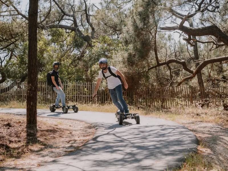 Deux hommes portant casque et sac à dos pratiquent le skate électrique sur un sentier sinueux en forêt.