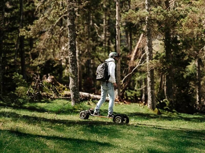 Homme avec sac à dos et casque utilise un skate électrique sur une surface herbeuse lors d'une balade dans le bois de Boulogne.