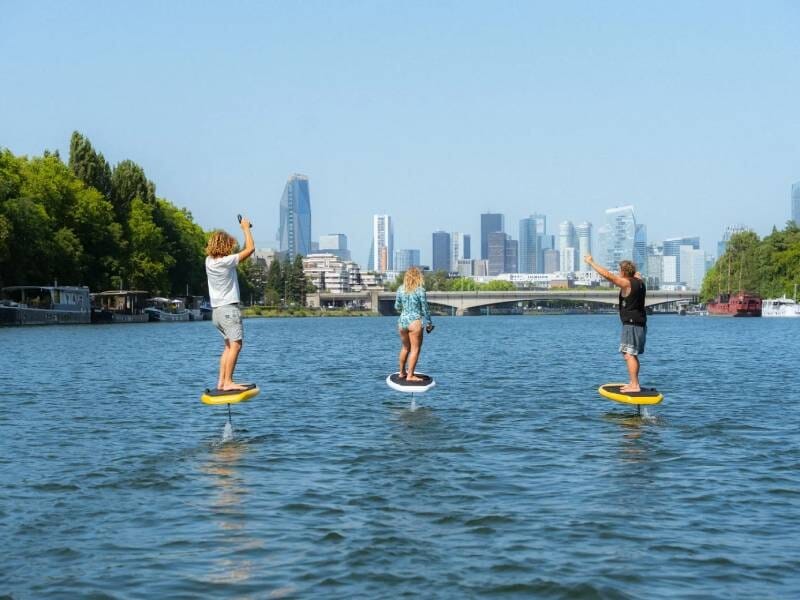 Trois personnes pratiquent le surf électrique sur la Seine avec la ville de Paris en arrière-plan sous un ciel bleu.