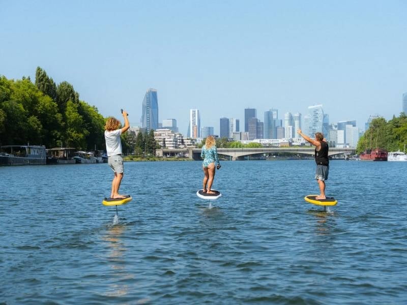Trois personnes pratiquent le surf électrique sur la Seine avec la ville de Paris en arrière-plan sous un ciel bleu.