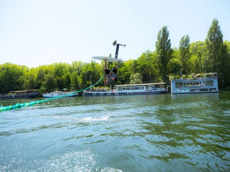 Un homme en train de faire du surf électrique en sautant au-dessus de l'eau sur la Seine avec des arbres et des bateaux.