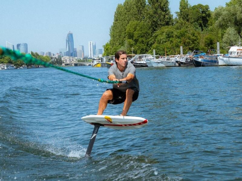 Un homme sur un surf électrique tracté par une corde sur la Seine, avec des bateaux et des arbres en arrière-plan.