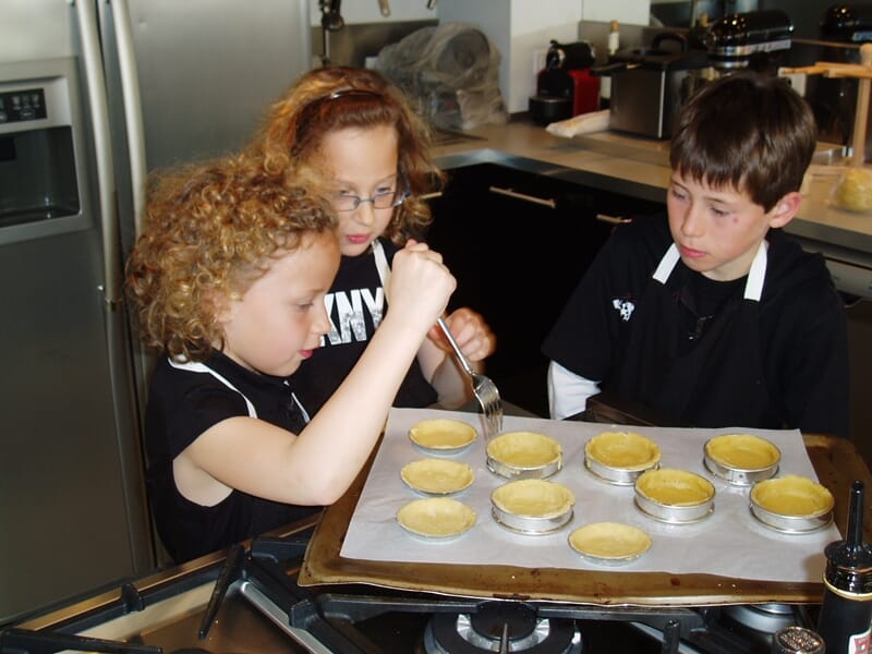 Trois enfants concentrés en tabliers noirs préparent des tartes lors d'un cours de cuisine à Nice. 