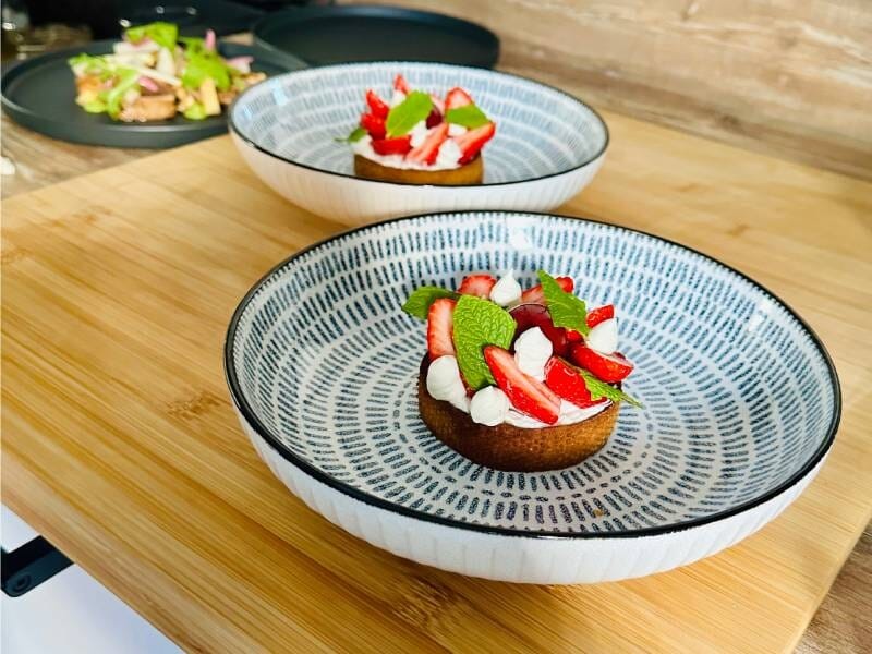Deux desserts élégants avec fraises, kiwi et crème sur assiettes décorées posées sur une table en bois clair.
