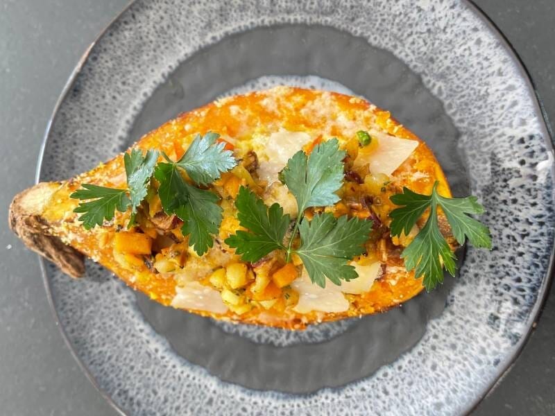 Plat principal avec une patate douce garnie de légumes, herbes fraîches et sauce, présenté sur une assiette grise.