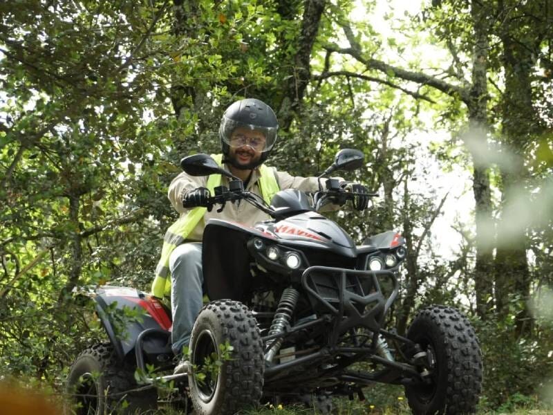 Homme en gilet jaune conduisant un quad noir et rouge dans un sentier forestier verdoyant sous un ciel clair.