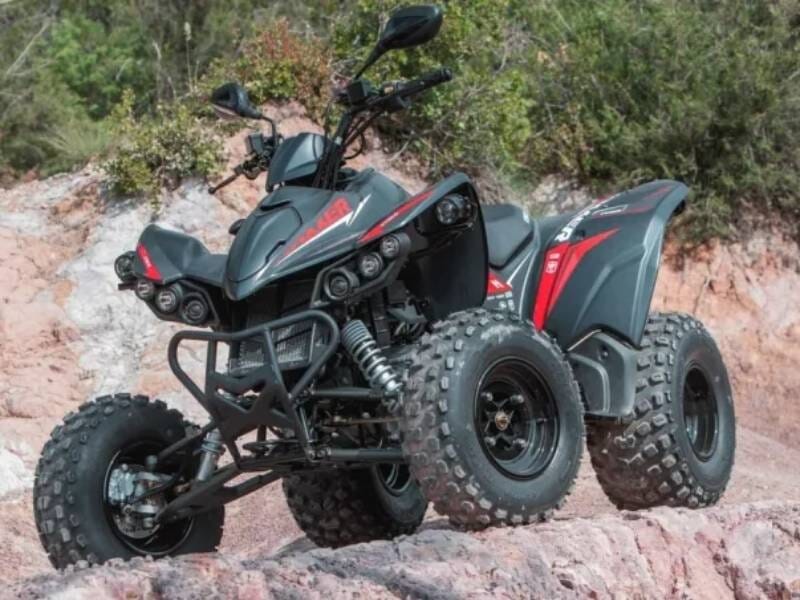 Quad noir et rouge garé sur un terrain rocheux avec des buissons verts en arrière-plan sous un ciel nuageux.