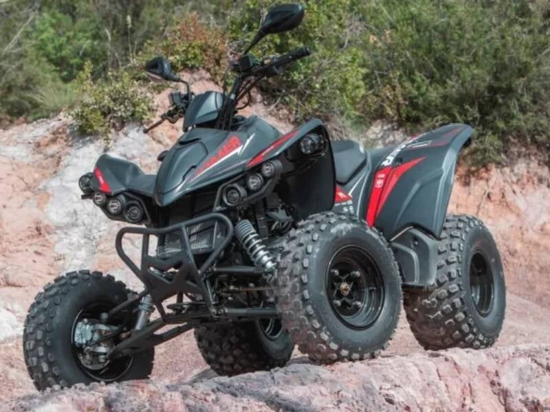 Quad noir et rouge garé sur un terrain rocheux avec des buissons verts en arrière-plan sous un ciel nuageux.