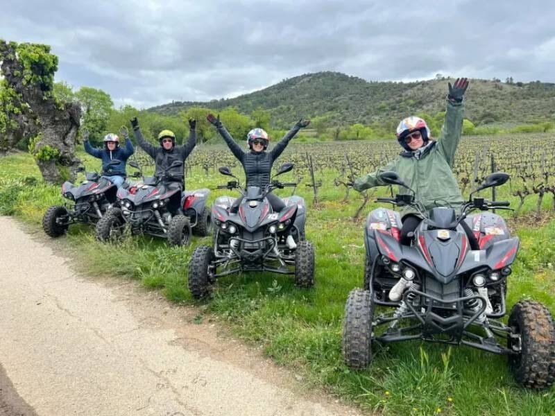 Quatre personnes en tenue de protection posant sur leurs quads noirs et rouges sur un chemin entouré de vignes, lors d'une randonnée à Vallon-Pont-d'Arc.