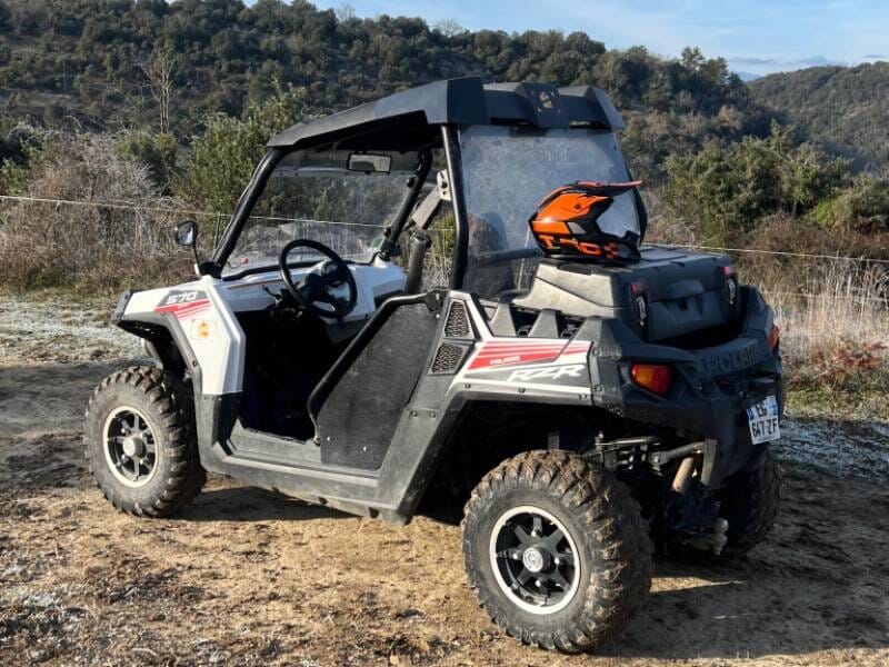 Quad tout-terrain blanc garé sur un chemin de terre avec un casque orange posé sur le toit, paysage naturel en arrière-plan.