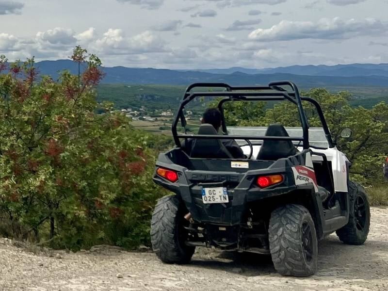 Vue arrière d'un quad blanc sur un chemin de terre avec un conducteur, paysage vallonné et ciel nuageux en arrière-plan.