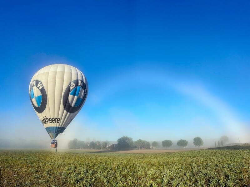 Montgolfière blanche avec logo BMW survolant un champ vert lors d'un vol en montgolfière près de la Montagne de Lure.