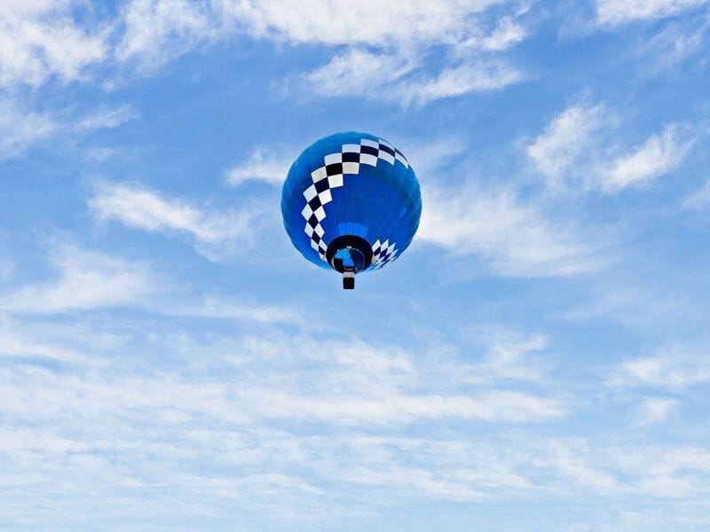Montgolfière bleue avec motifs blancs volant dans un ciel clair parsemé de nuages légers.