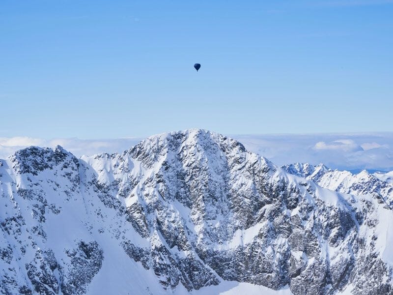 Montgolfière lointaine volant au-dessus de montagnes enneigées sous un ciel bleu clair.