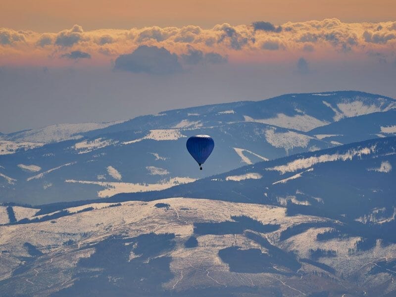 Montgolfière bleue survolant des montagnes avec des zones enneigées au coucher du soleil.