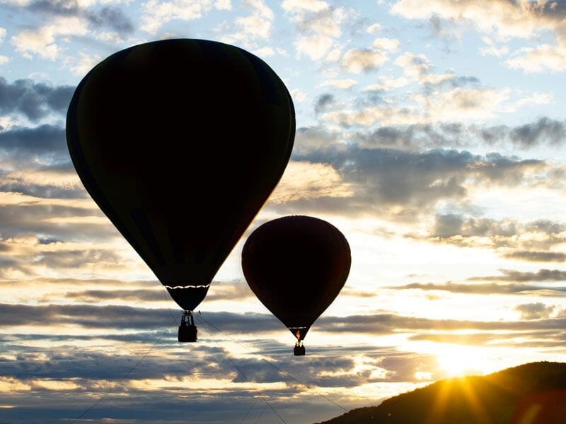 Deux montgolfières en silhouette au coucher du soleil avec un ciel nuageux et le soleil bas.