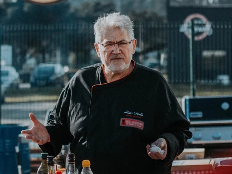 Homme âgé en veste de chef explique une recette lors d'un atelier de cuisine barbecue à Brest.