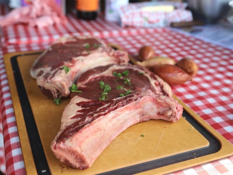 Deux grosses côtes de viande crue avec un peu de persil sur une planche, sur une table à carreaux rouges et blancs.