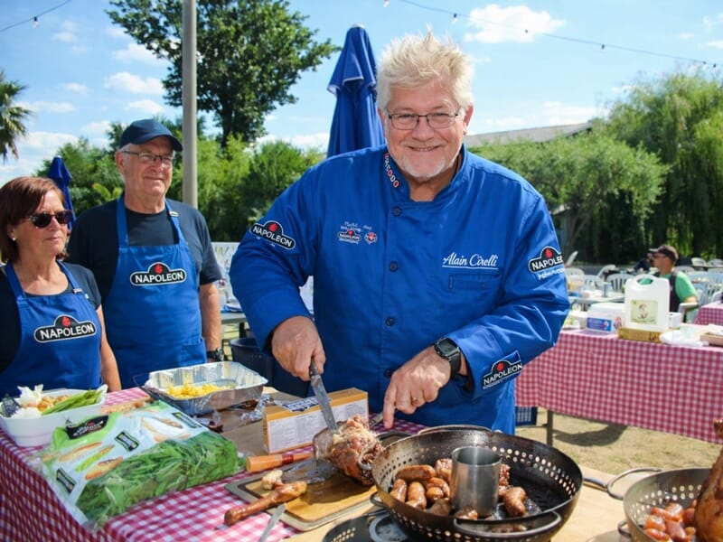 Un homme souriant en veste bleue découpe de la viande lors d'un cours de cuisine en plein air avec d'autres participants.