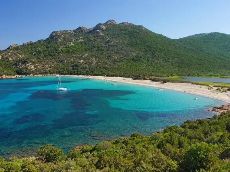 Vue aérienne d'une plage de sable blanc avec une mer turquoise et un bateau à voile ancré près du rivage, lors d'une location de bateau à Santa-Giulia.