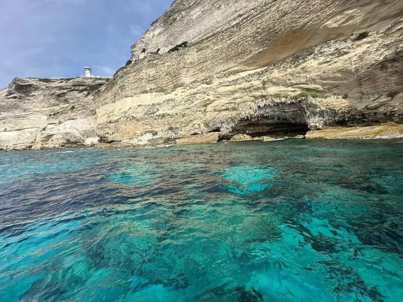 Eaux cristallines turquoise au pied d'une falaise rocheuse beige avec une petite grotte visible dans la roche, lors d'une balade en bateau à Santa-Giulia.