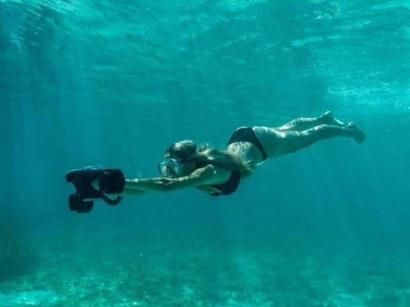 Femme en maillot de bain nageant sous l'eau avec un scooter de plongée électrique dans un environnement marin transparent.