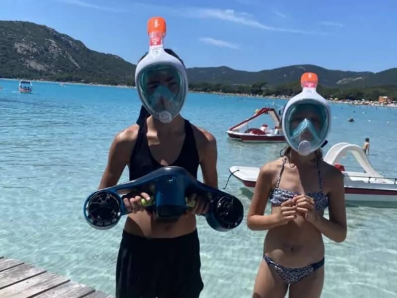 Deux enfants en maillot de bain avec masque de plongée intégral, tenant un scooter sous-marin sur une plage au bord d'une mer turquoise, près de Porto-Vecchio.