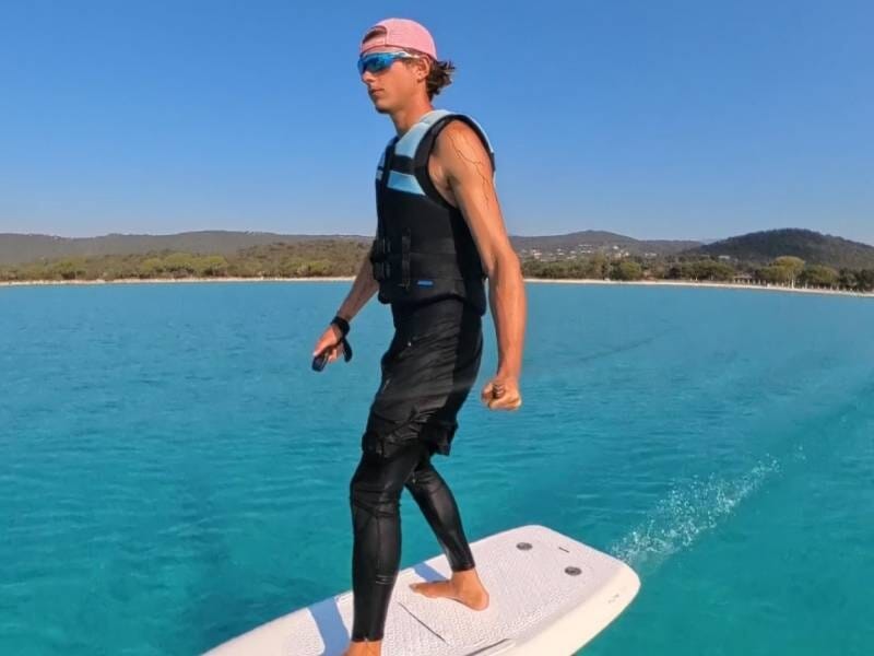Jeune homme en combinaison et casquette rose sur un surf électrique glissant sur l'eau claire à Santa Giulia.