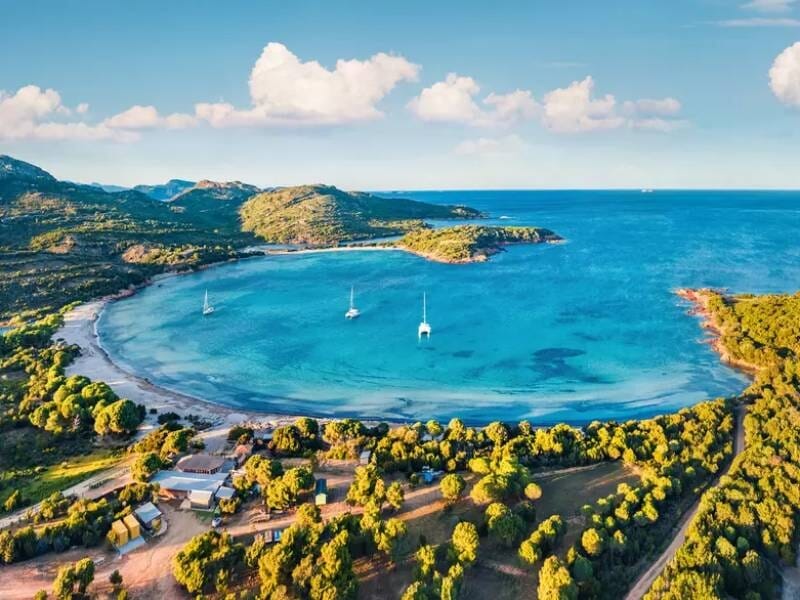 Vue aérienne d'une baie turquoise avec plusieurs voiliers ancrés près d'une plage bordée de végétation dense et collines