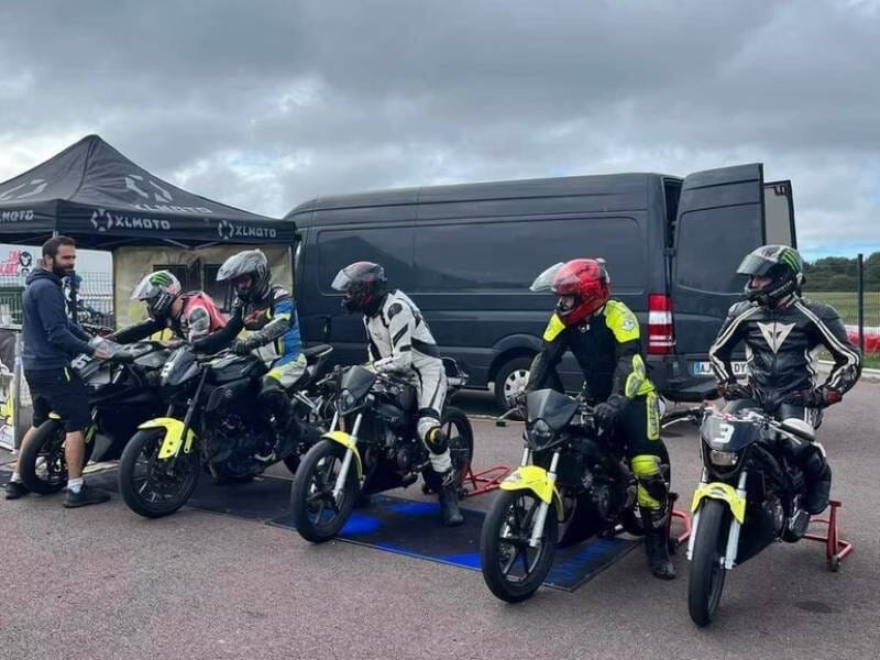 Quatre pilotes en tenue de moto et casques alignés près de leurs motos sous un ciel nuageux sur une piste.