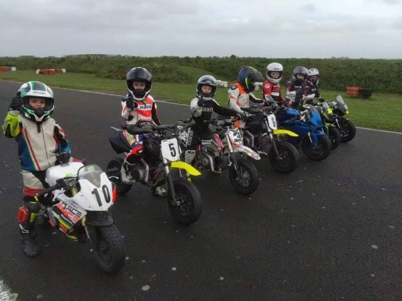 Six enfants en tenue de moto posent sur leurs petites motos sur une piste de course sous un ciel nuageux.
