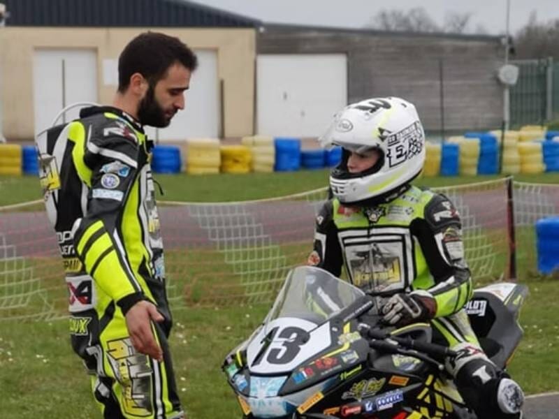 Un instructeur en combinaison discute avec un jeune pilote casqué sur une moto de course sur circuit.