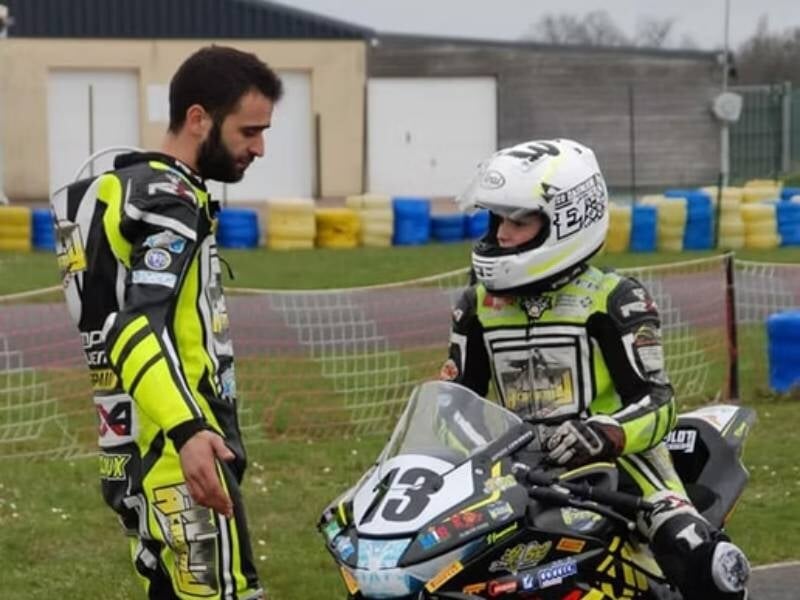 Un instructeur en combinaison discute avec un jeune pilote casqué sur une moto de course sur circuit.