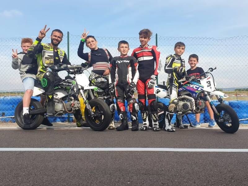 Groupe de jeunes pilotes en combinaison posant joyeusement avec leurs motos sur une piste de course lors d'un stage de pilotage au circuit karting de Lessay.