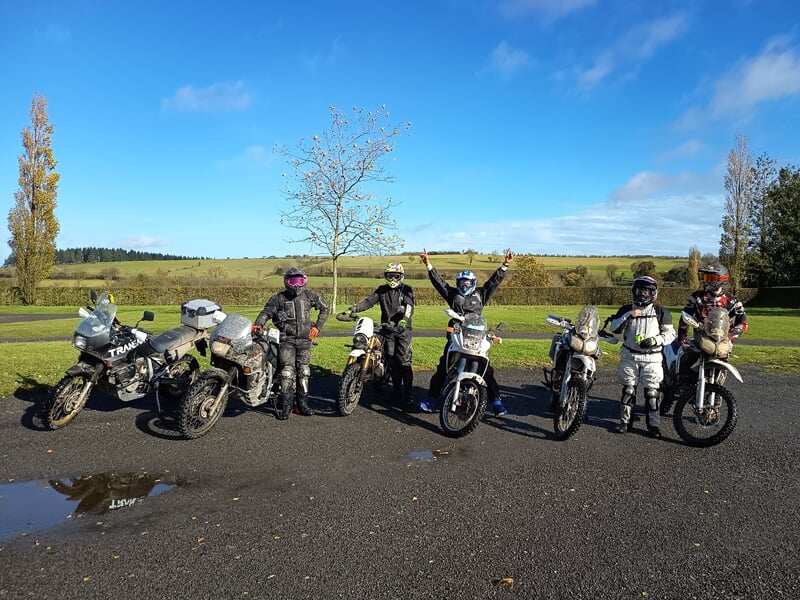 Groupe de six motards posant avec leurs motos sur une route goudronnée sous un ciel bleu clair et un paysage champêtre.