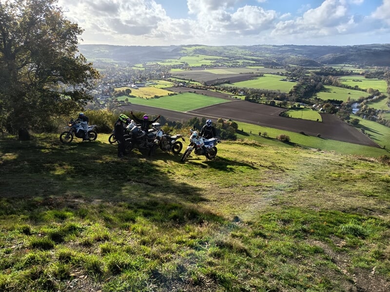 Cinq motards avec leurs motos tout-terrain sur une colline offrant une vue panoramique sur une vallée agricole verdoyante.