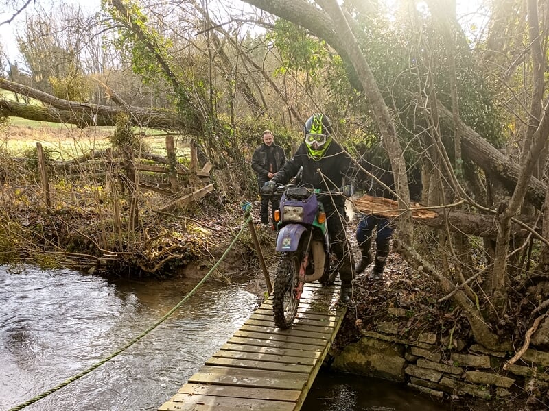 Motard franchissant un petit pont en bois au-dessus d'un ruisseau, aidé par une personne dans un environnement boisé.